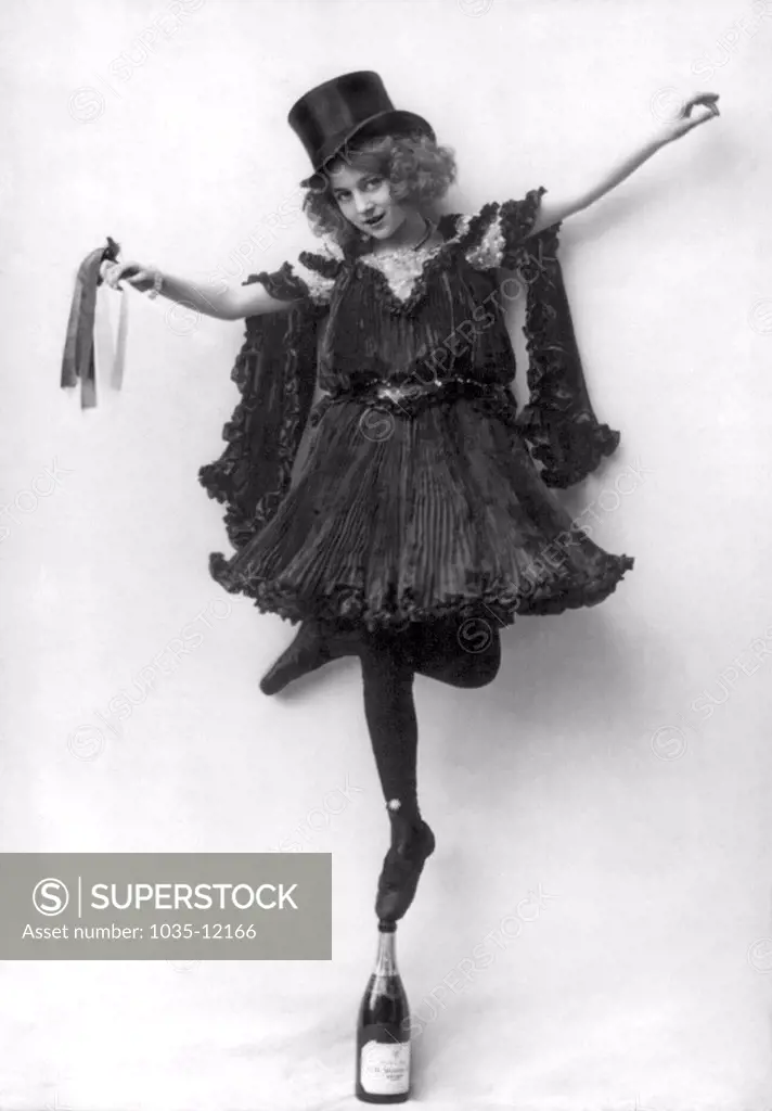 Providence, Rhode Island:  1904. A woman in an ornate costume with black pressed pleats and a top hat, standing en pointe on a champagne bottle.