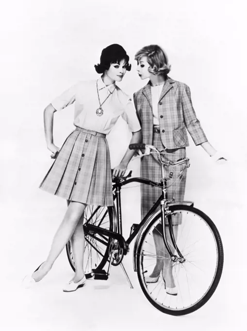 United States:  c. 1958 Two women with a bicycle model vacation fashions in cotton for traveling or sightseeing. They are styled by Miss Pat of California.