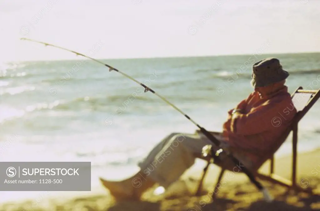 Senior man sitting on the beach on a deck chair with a fishing rod