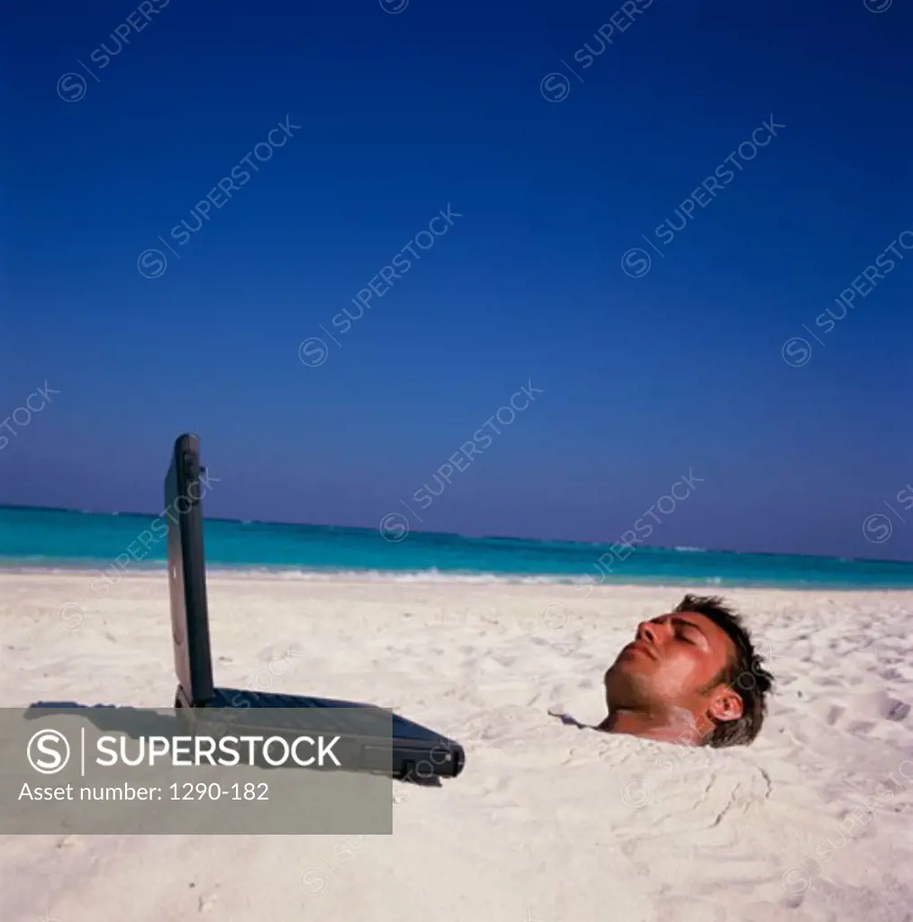 Young man buried in sand on the beach with a laptop in front of him