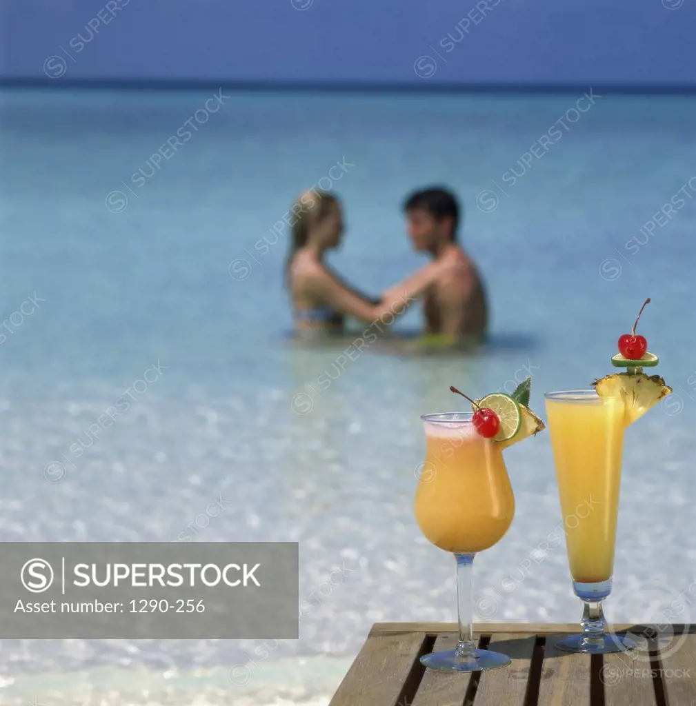 Two cocktail drinks on a table at the beach with a young couple behind in  the water - SuperStock