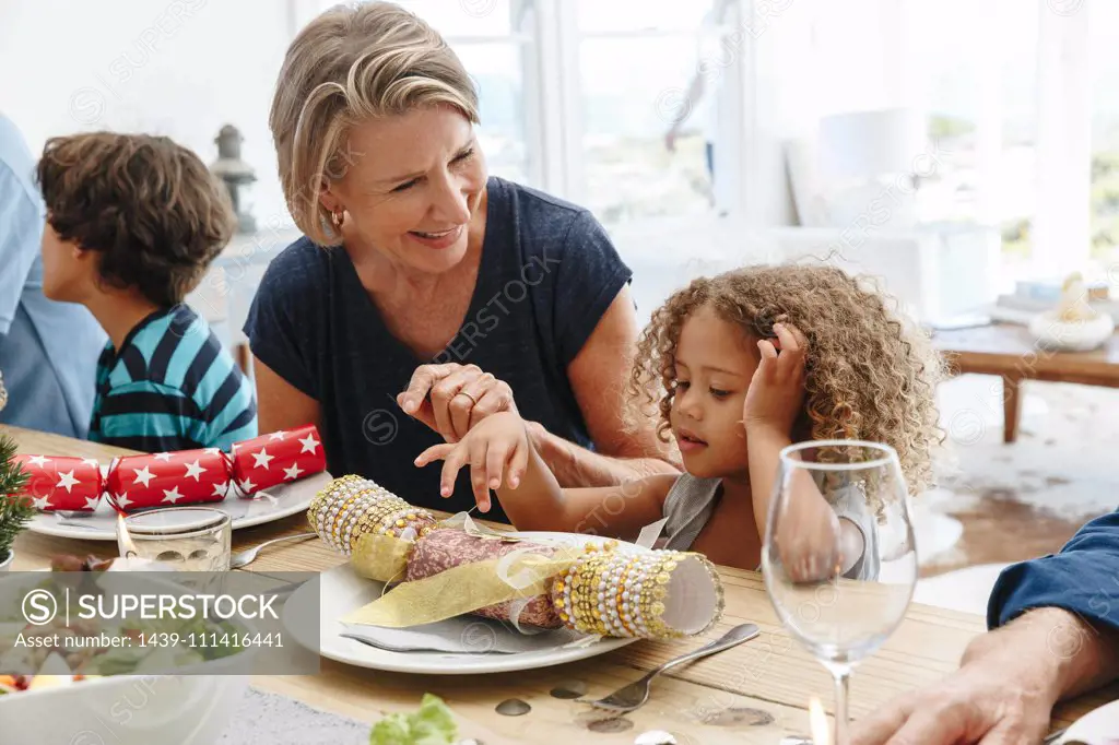 Woman talking to granddaughter playing with Christmas cracker at dining table