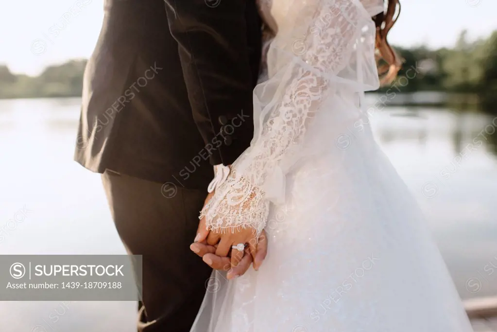 Mid section of bride and groom holding hands on lakeshore