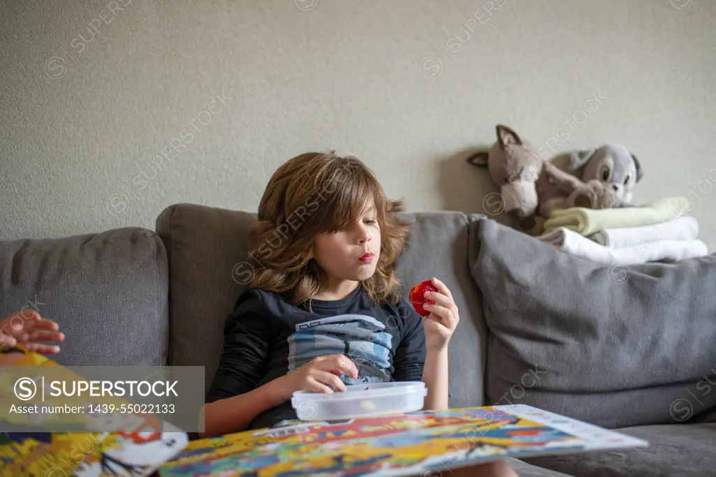 Young boy sitting on the sofa eating healthy strawberries and reading a book