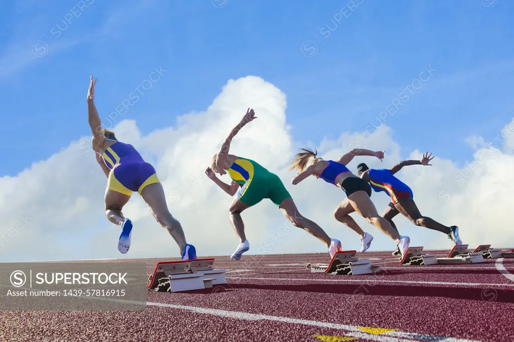 Four female athletes on athletics track, leaving starting blocks, rear view