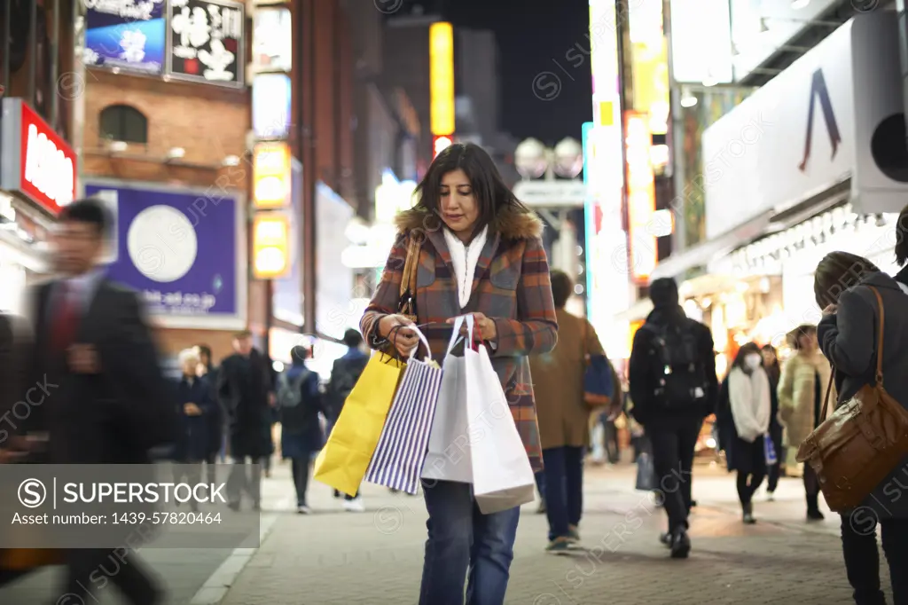 Front view of mature woman carrying shopping bags in city at night looking down, Shibuya, Tokyo, Japan