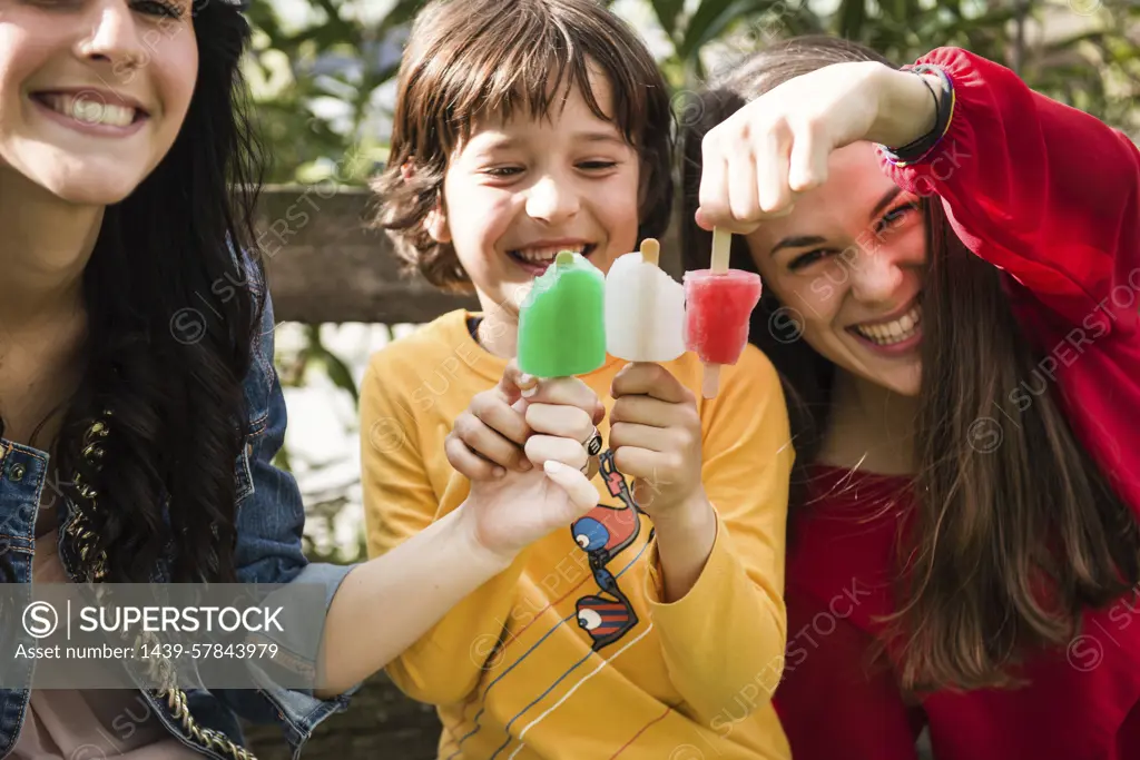 Two young women and young boy, sitting on bench, holding ice lollies together to create colours of Italian flag