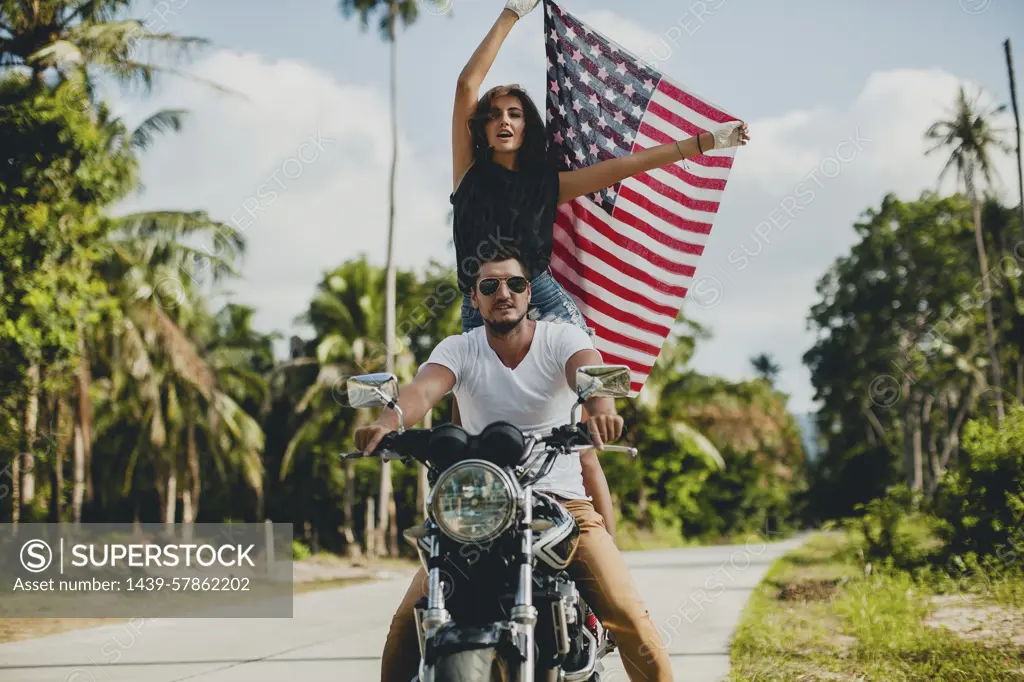 Young couple holding up American flag while riding motorcycle on rural road, Krabi, Thailand