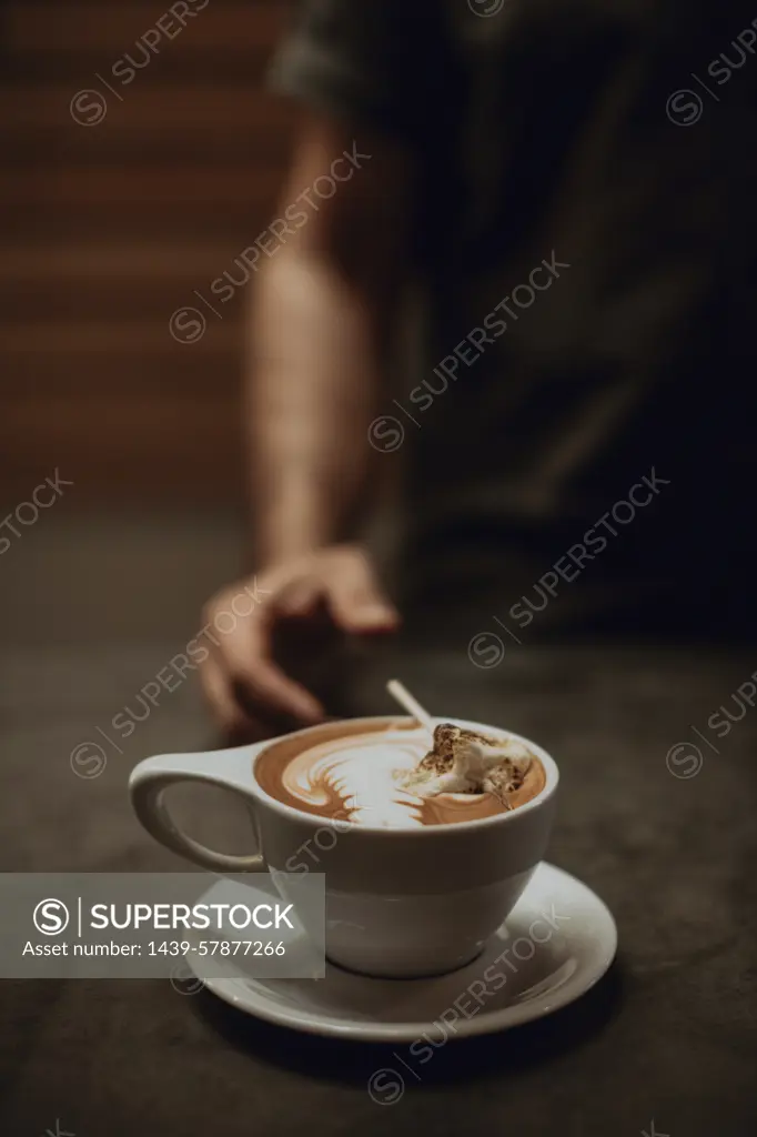 Barista at cafe counter with cup of mocha topped with toasted marshmallow, cropped shallow focus