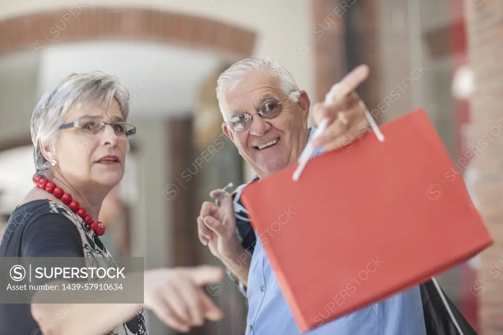 Mature couple walking along street, carrying shopping bags, pointing behind them