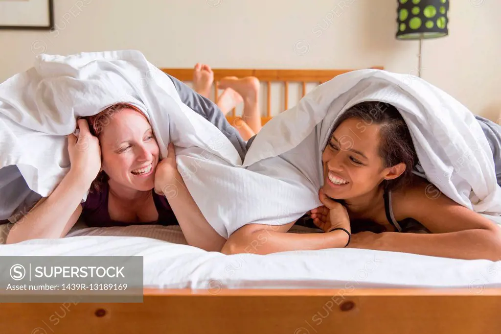 Young women lying on front in bed underneath quilt face to face smiling