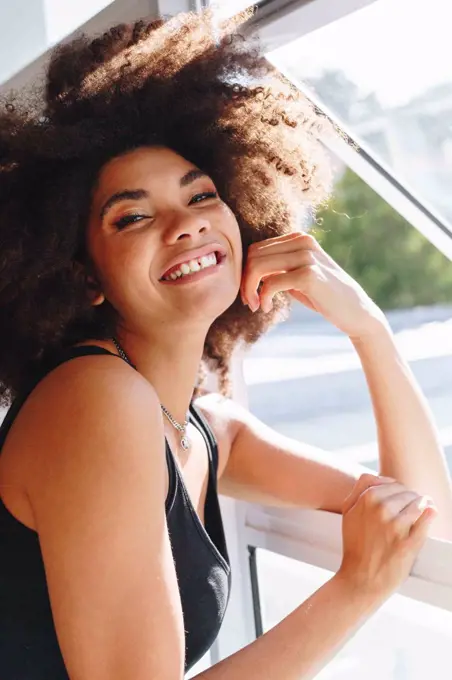 Happy young woman with afro hairstyle at bedroom window, head and shoulder portrait