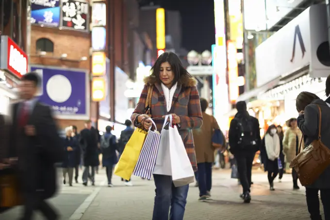 Front view of mature woman carrying shopping bags in city at night looking down, Shibuya, Tokyo, Japan