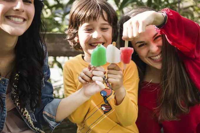 Two young women and young boy, sitting on bench, holding ice lollies together to create colours of Italian flag