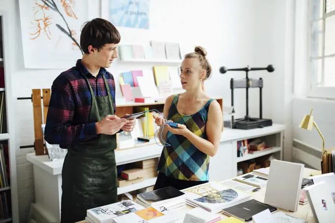 Young woman chatting to young male craftsman and using tablet technology to make purchase in art bookshop