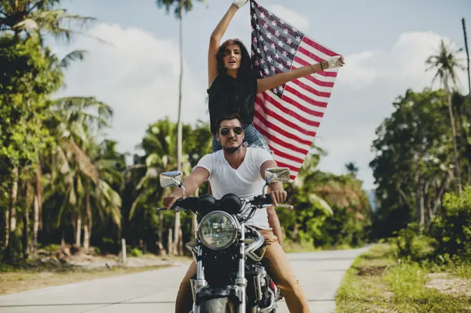 Young couple holding up American flag while riding motorcycle on rural road, Krabi, Thailand