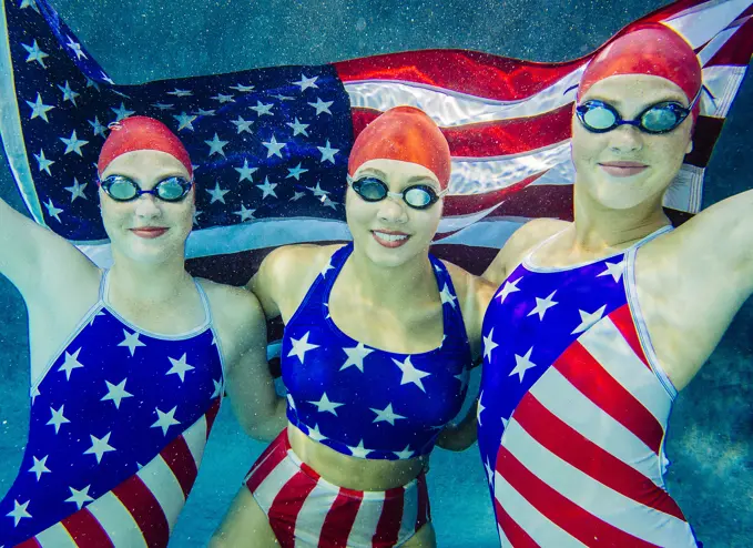 Three female swimmers, underwater, wearing stars and stripes costumes, holding american flag