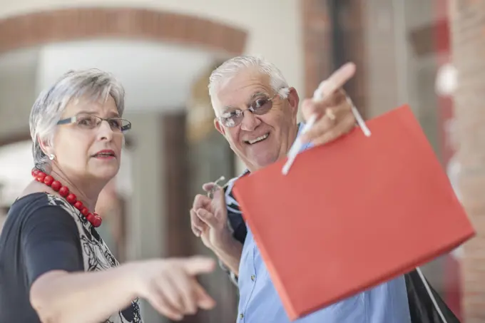 Mature couple walking along street, carrying shopping bags, pointing behind them