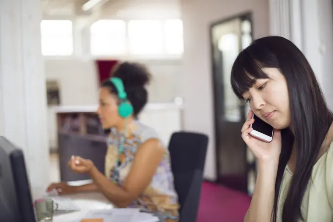 Two women working in an office environment, one talking on the phone and the other focused on her computer monitor.