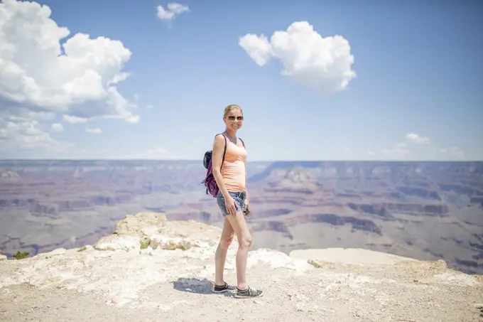 Smiling woman with sunglasses and backpack standing at the edge of the Grand Canyon on a sunny day.