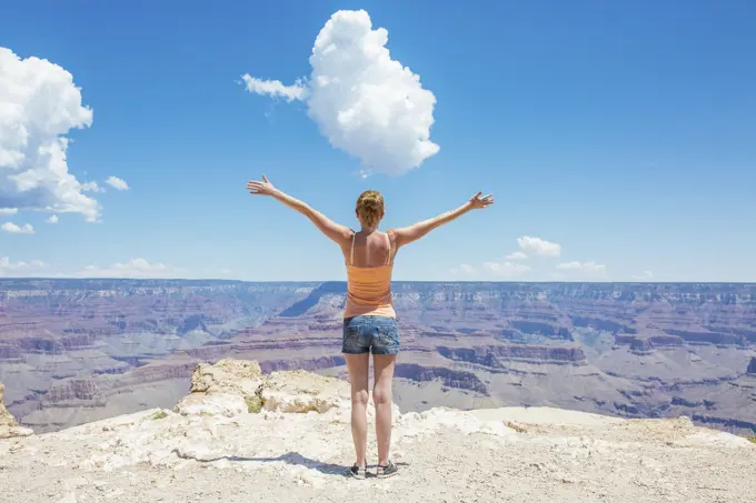 Woman with arms spread wide standing at the edge of the Grand Canyon under a blue sky.