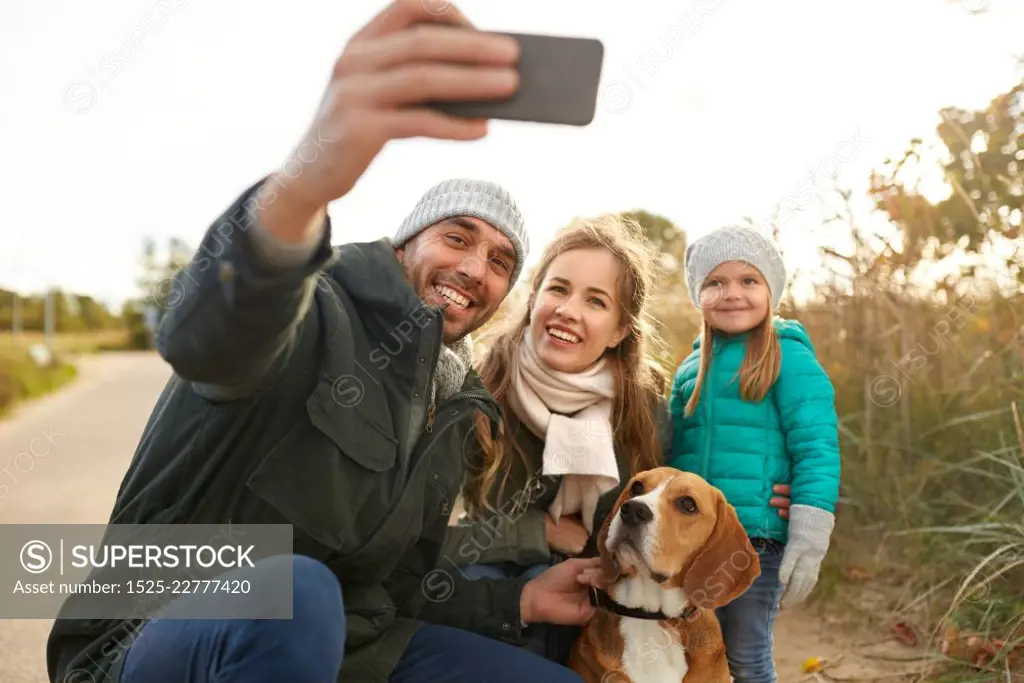 family, pets and people concept - happy mother, father and little daughter with beagle dog taking selfie by smartphone outdoors in autumn. happy family with dog taking selfie in autumn