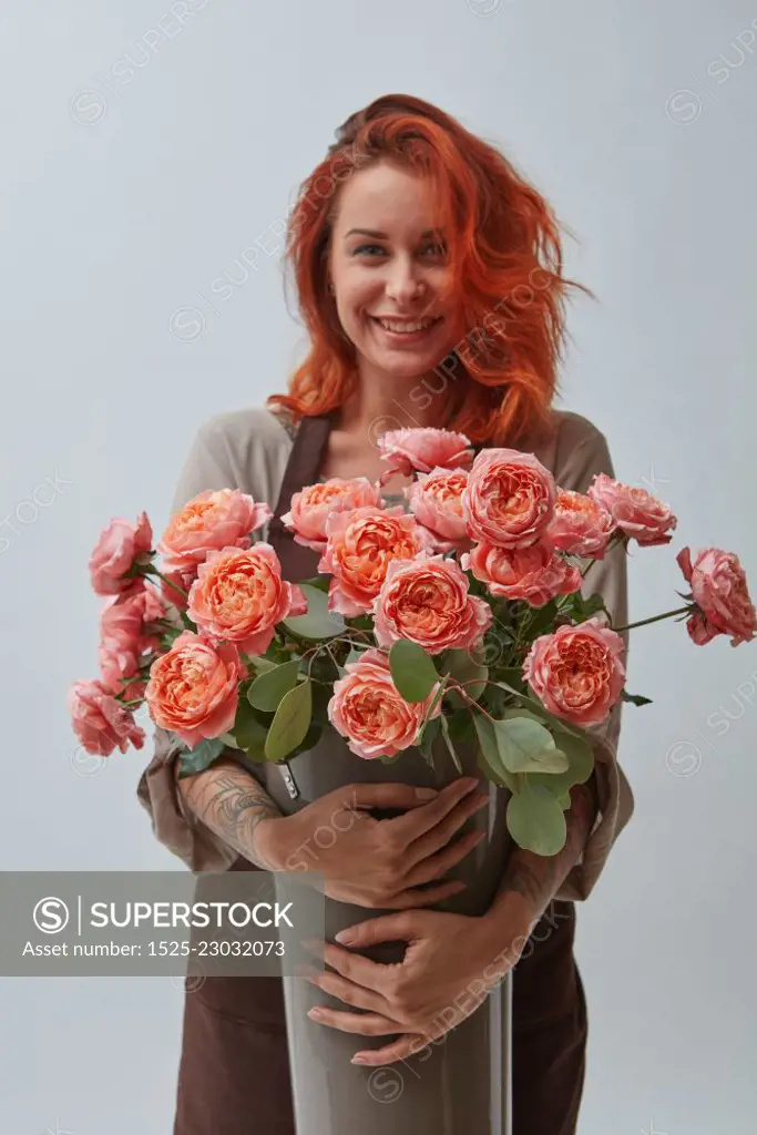 A smiling red-haired woman in a brown apron holds a vase with roses in a color of the year 2019 Living Coral Pantone on a gray background with space for text. The concept of a flower shop. Young woman with a big vase of roses flowers in a color of the year 2019 Living Coral Pantone on a gray background with copy space.