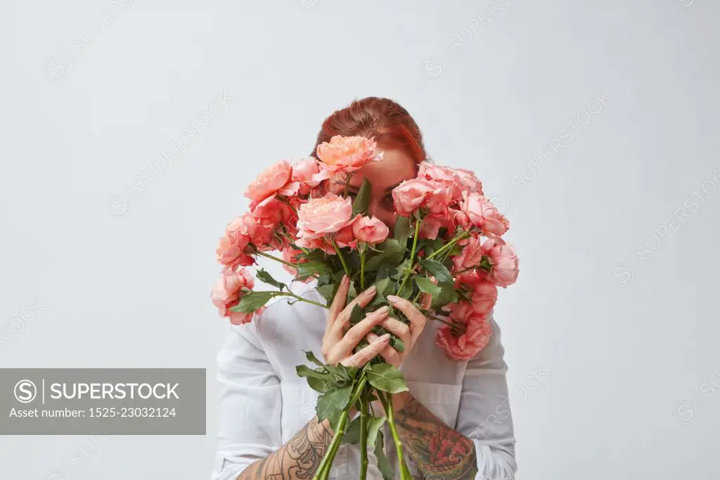 A happy woman with a tattoo on her hands hides her face behind a bouquet of fresh roses in a color of the year 2019 Living Coral Pantone. Postcards for Mother&rsquo;s Day.. Congratulation bouquet of roses in the gerl&rsquo;s hands with a tattoo on a white background. Trend color of the year 2019 Living Coral Pantone.