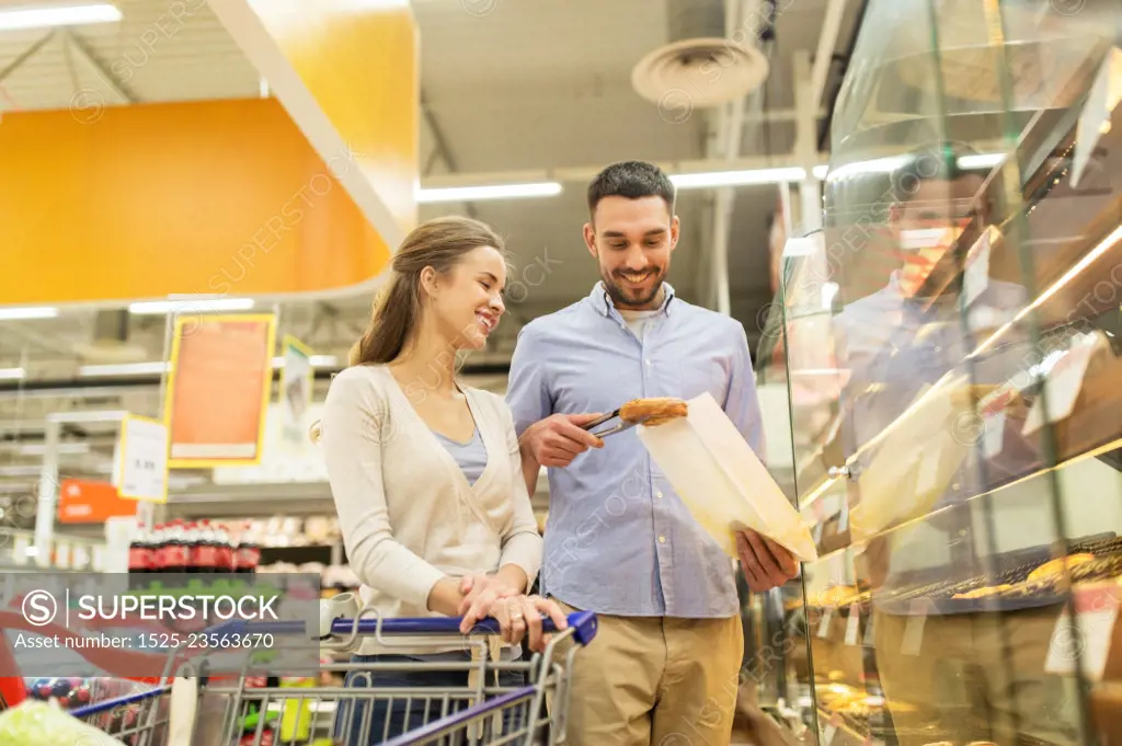 food, sale, consumerism and people concept - happy couple with shopping cart at grocery store or supermarket baking department buying buns or pies. happy couple with shopping cart at grocery store