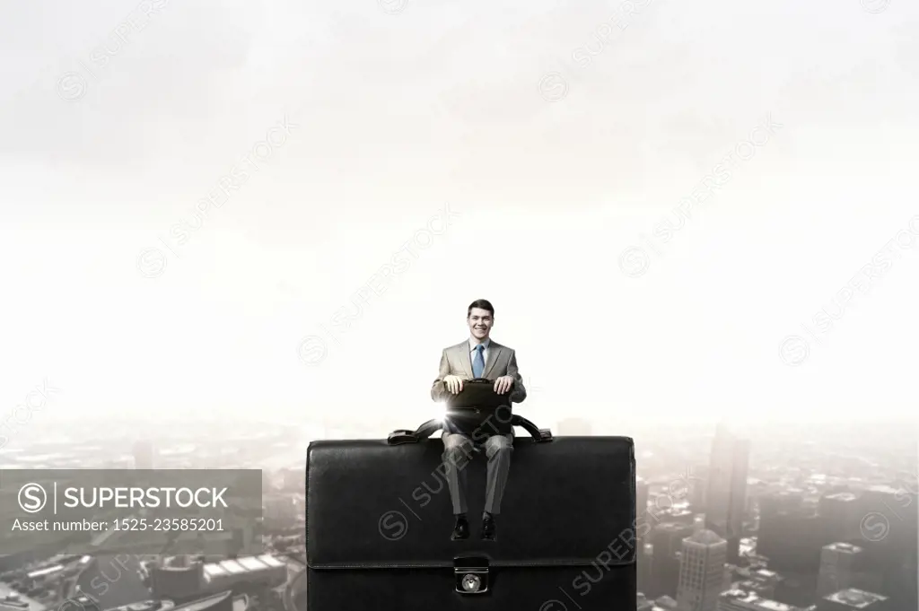 Businessman on big suitcase. Young smiling businessman sitting on giant briefcase