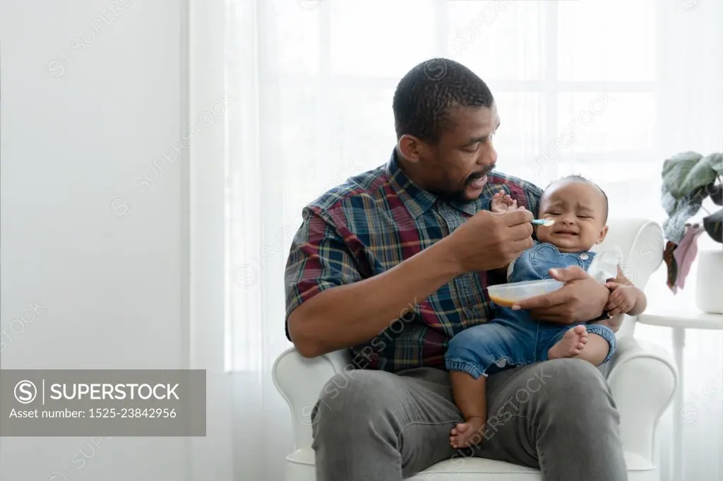 African toddler baby girl crying and dont want to eat food from her father who feeding with spoon while sitting on sofa at home. Little child care and relationship of dad and little daughter concept