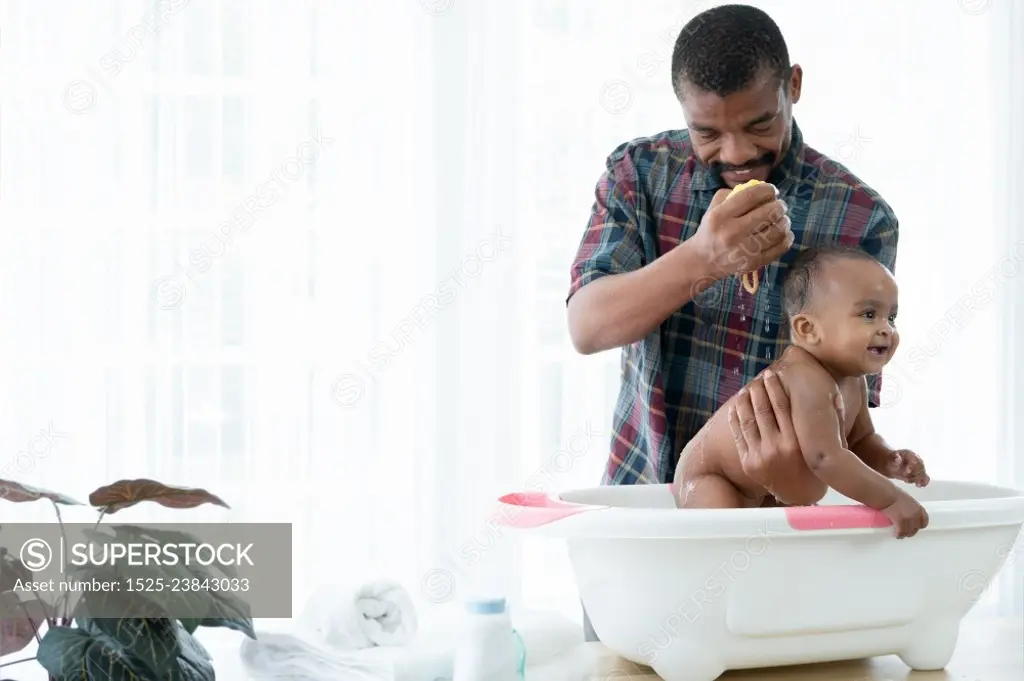 Happy African father with beard bathing adorable newborn baby daughter in bathtub at home. Dad cleaning his little kid girl with sponge in warm water. Child hygiene cleanliness care concept