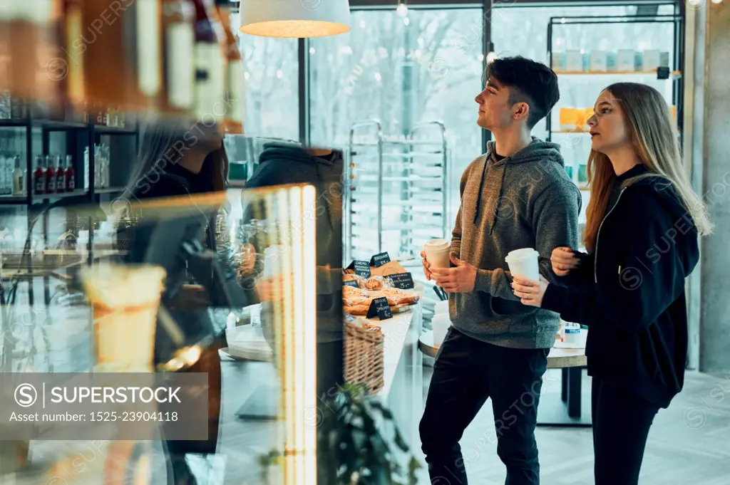 Friends doing shopping in a coffee shop. Young man and woman having chat while picking out the pastry, bakery&rsquo;s goods and hot drinks standing at counter in a coffee shop. People buying coffee and sweet snacks to go