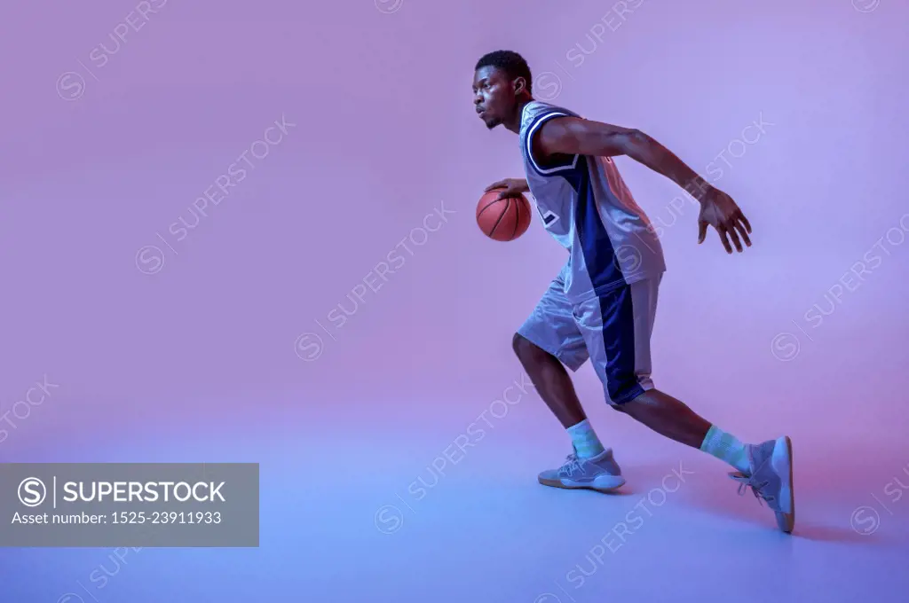 Basketball player practicing with ball in studio, neon background. Professional male baller in sportswear playing sport game, tall sportsman. Basketball player practicing with ball in studio