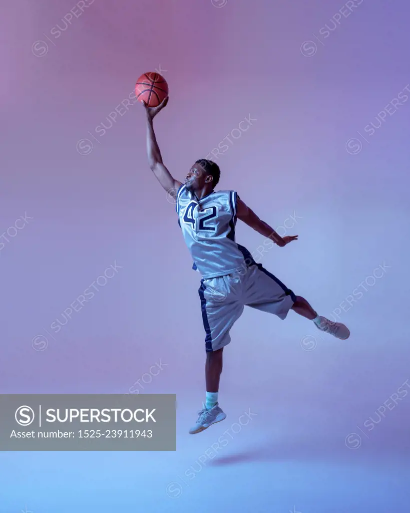 Basketball player with ball shows his skill in studio, jump in action, neon background. Professional male baller in sportswear playing sport game, tall sportsman. Basketball player with ball, jump in action