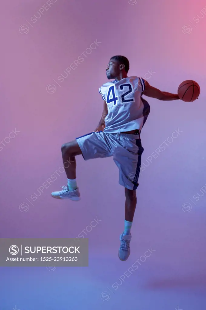 Basketball player jumping with ball in studio, neon background. Professional male baller in sportswear playing sport game. Basketball player jumping with ball in studio