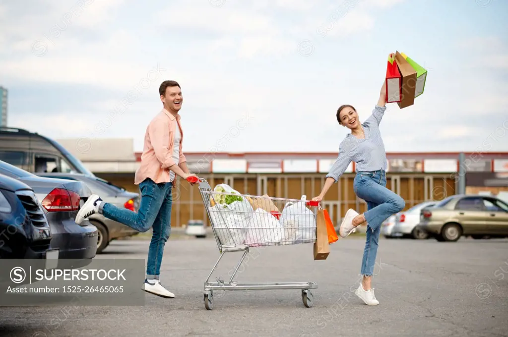 Cheerful family couple with bags in cart on supermarket car parking. Happy customers carrying purchases from the shopping center, vehicles on background. Cheerful family couple with cart on car parking