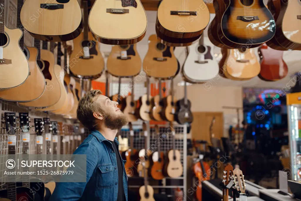 Bearded young guy choosing acoustic guitar in music store. Assortment in musical instruments shop, male musician buying equipment. Young guy choosing acoustic guitar in music store