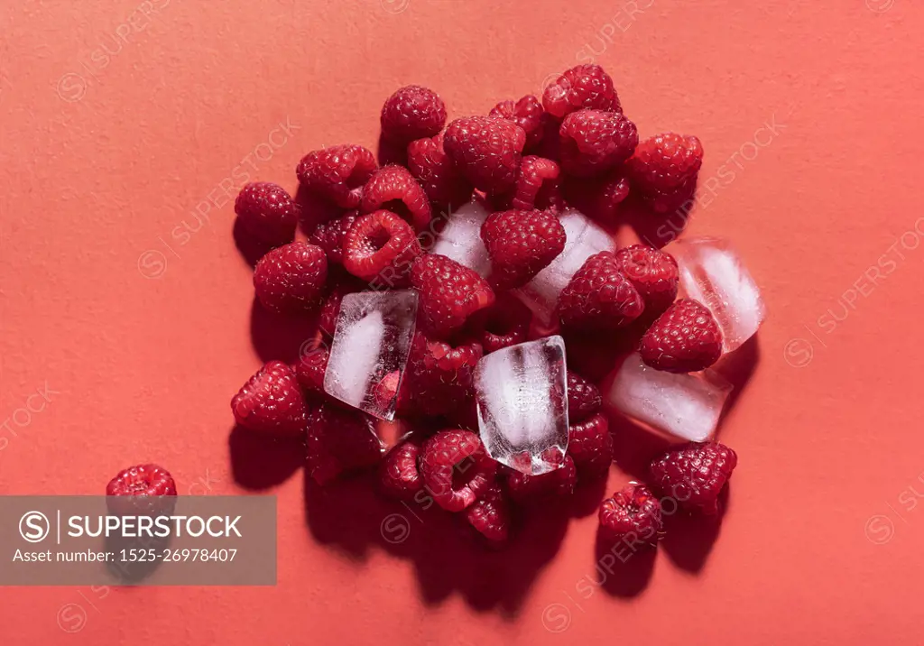 Fresh raspberries in a pile with ice cubes on a red background in sunlight. Above view of tasty summer fruits. Ingredients for berries lemonade.