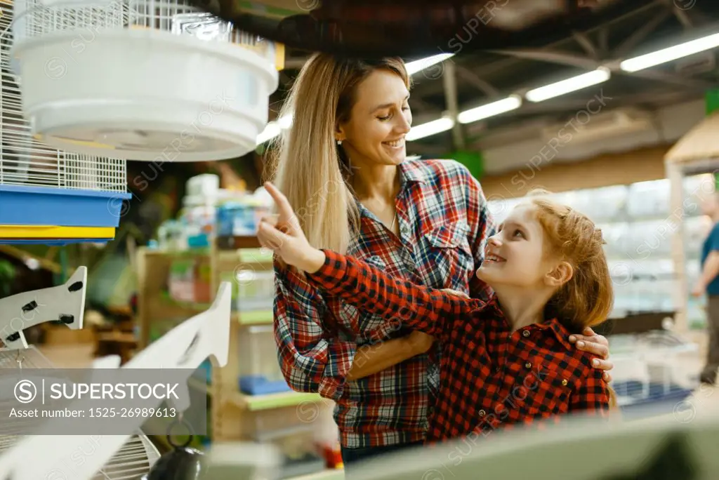 Mother with daughter choosing cage for bird at the showcase in pet store. Woman and little child buying equipment in petshop, accessories for domestic animals. Mother with daughter choosing cage in pet store