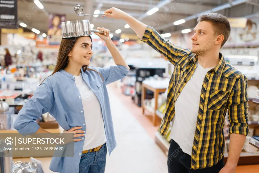 Young couple jokes with ladle and pan in houseware store. Man and woman buying home goods in market, family in kitchenware supply shop. Couple jokes with ladle and pan in houseware store