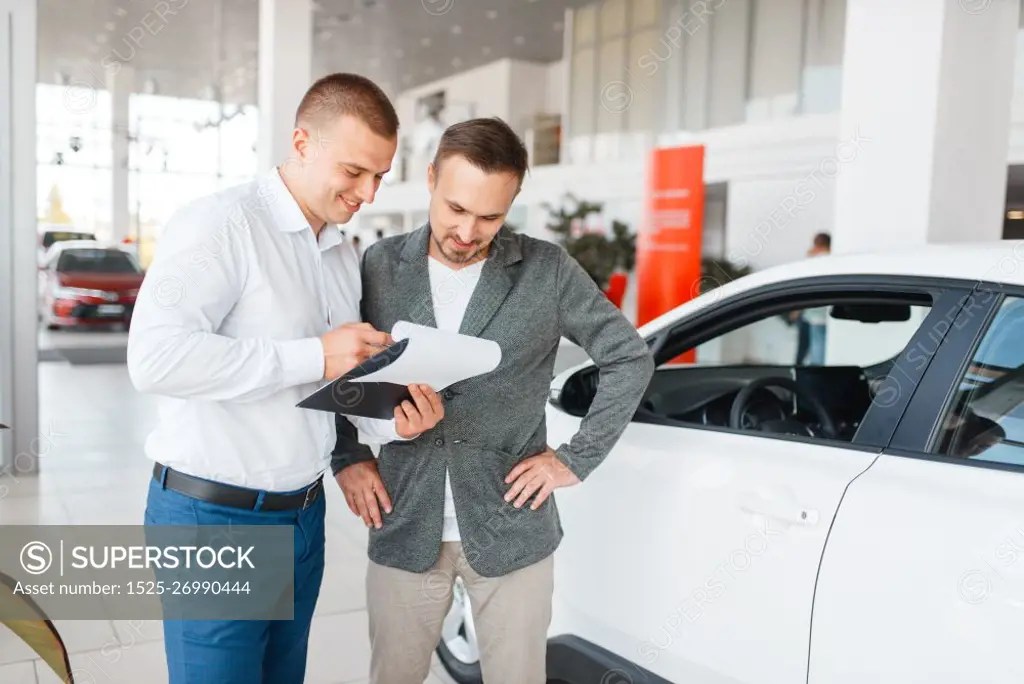 Salesman and buyer make out the purchase of new car in showroom. Male customer buying vehicle in dealership, automobile sale