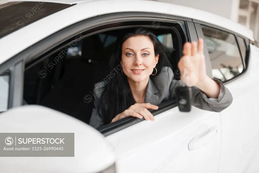 Happy woman shows the key to the new car in showroom. Female customer buying vehicle in dealership, automobile sale