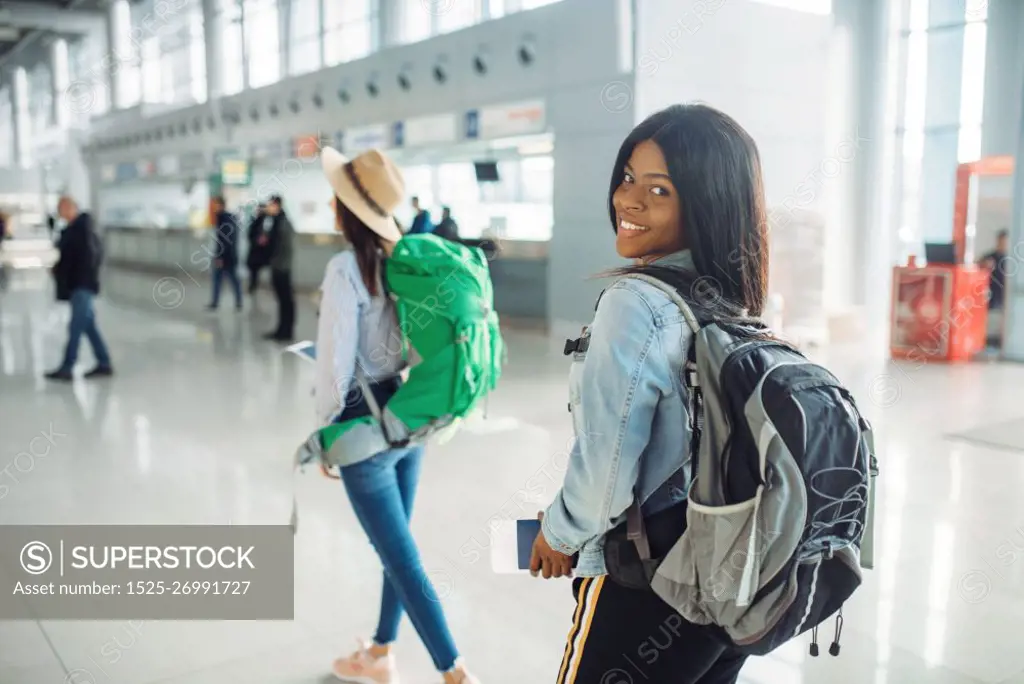 Two female tourists with backpacks in waiting area of international airport. Passengers with baggage in air terminal, back view, happy journey of white and black ladies, summer travel of happy women