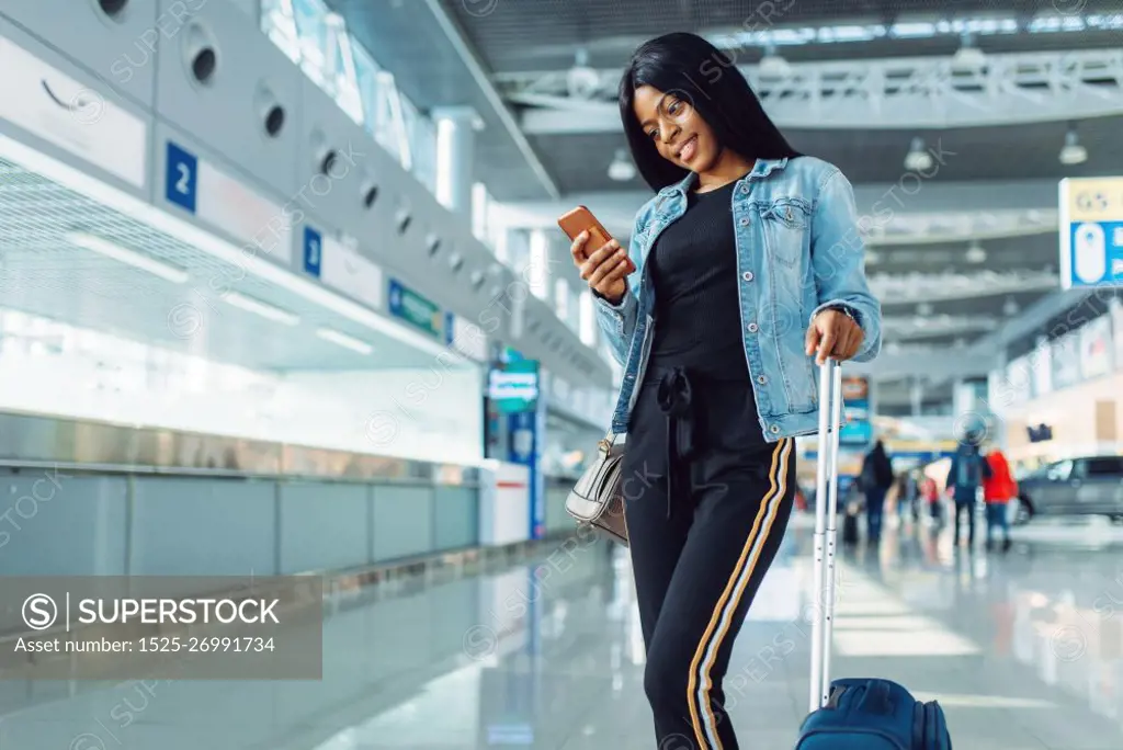 Black female tourist with suitcase and phone waiting for departure in international airport. Passenger with baggage in air terminal, happy journey sexy lady, summer travel of happy woman
