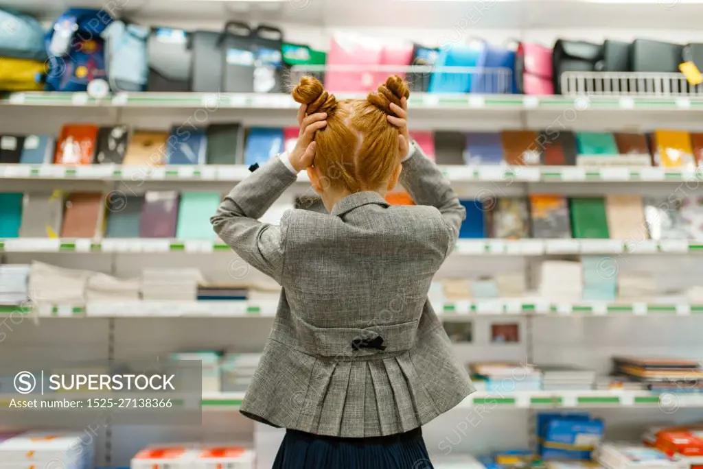 Little school girl at the shelf in stationery store, back view. Female child buying office supplies in shop, schoolchild in supermarket