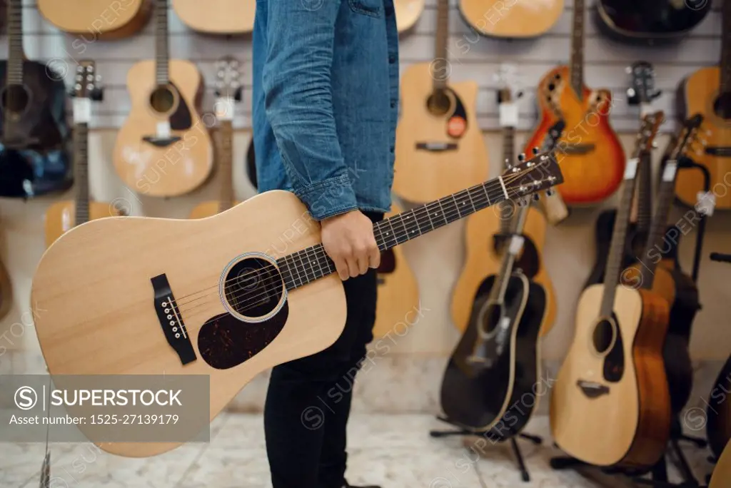 Young man holding acoustic guitar in music store. Assortment in musical instruments shop, male musician buying equipment