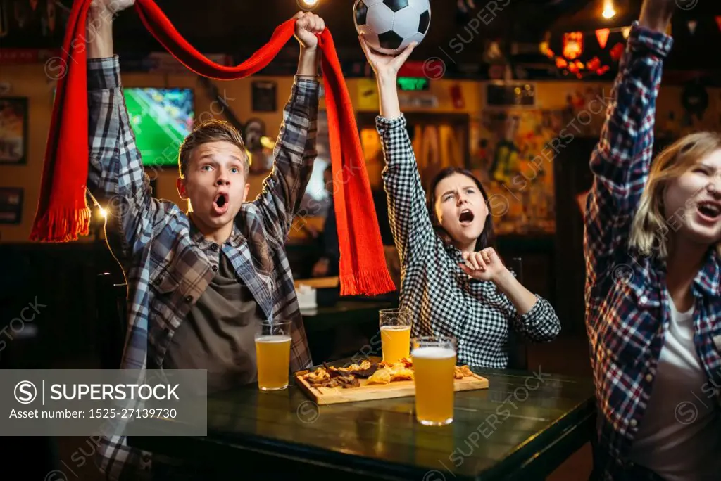 Football fans with scarf and ball in hands watching match and drinks beer in sports bar. Tv broadcasting, young friends leisures in pub, favorite team wins. Football fans with scarf and ball in sports bar