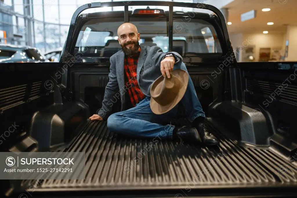 Smiling man poses in the back of new pickup truck in car dealership. Customer in vehicle showroom, male person buying transport, auto dealer business. Smiling man poses in the back of new pickup truck