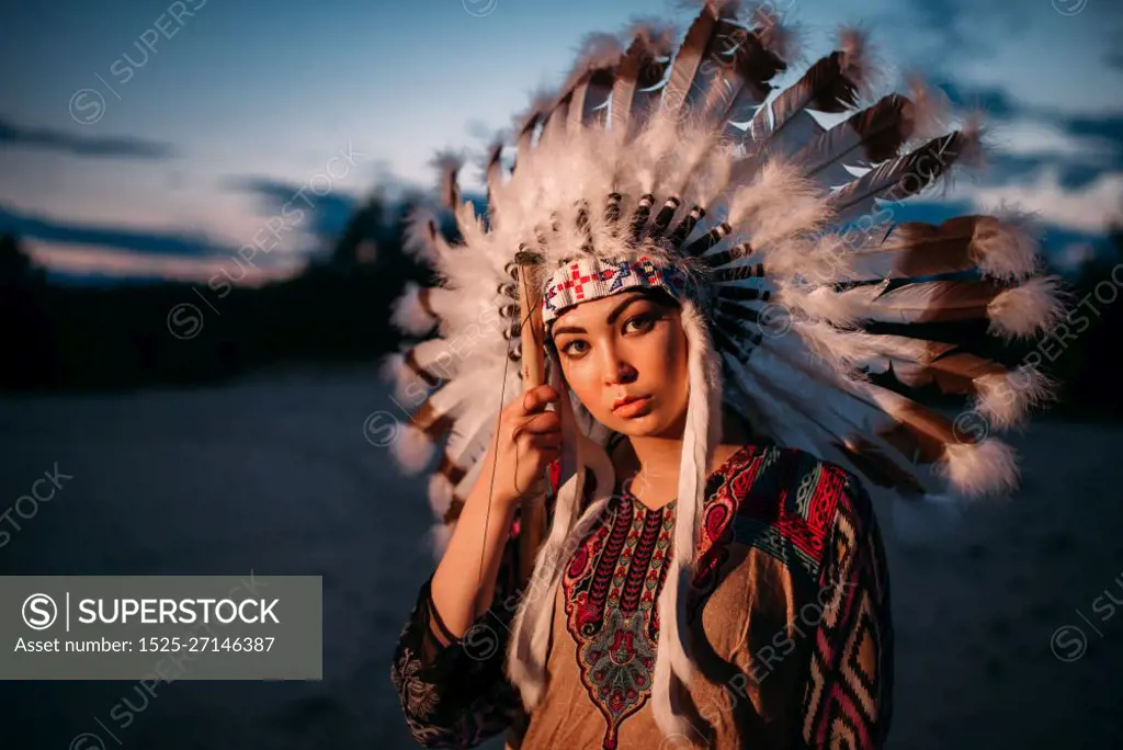 Portrait of young American Indian woman on sunset. Cherokee, Navajo, west native culture. Headdress made of feathers of wild birds. 